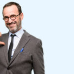 Teacher man using glasses proud, excited and arrogant, pointing with victory face isolated over blue background